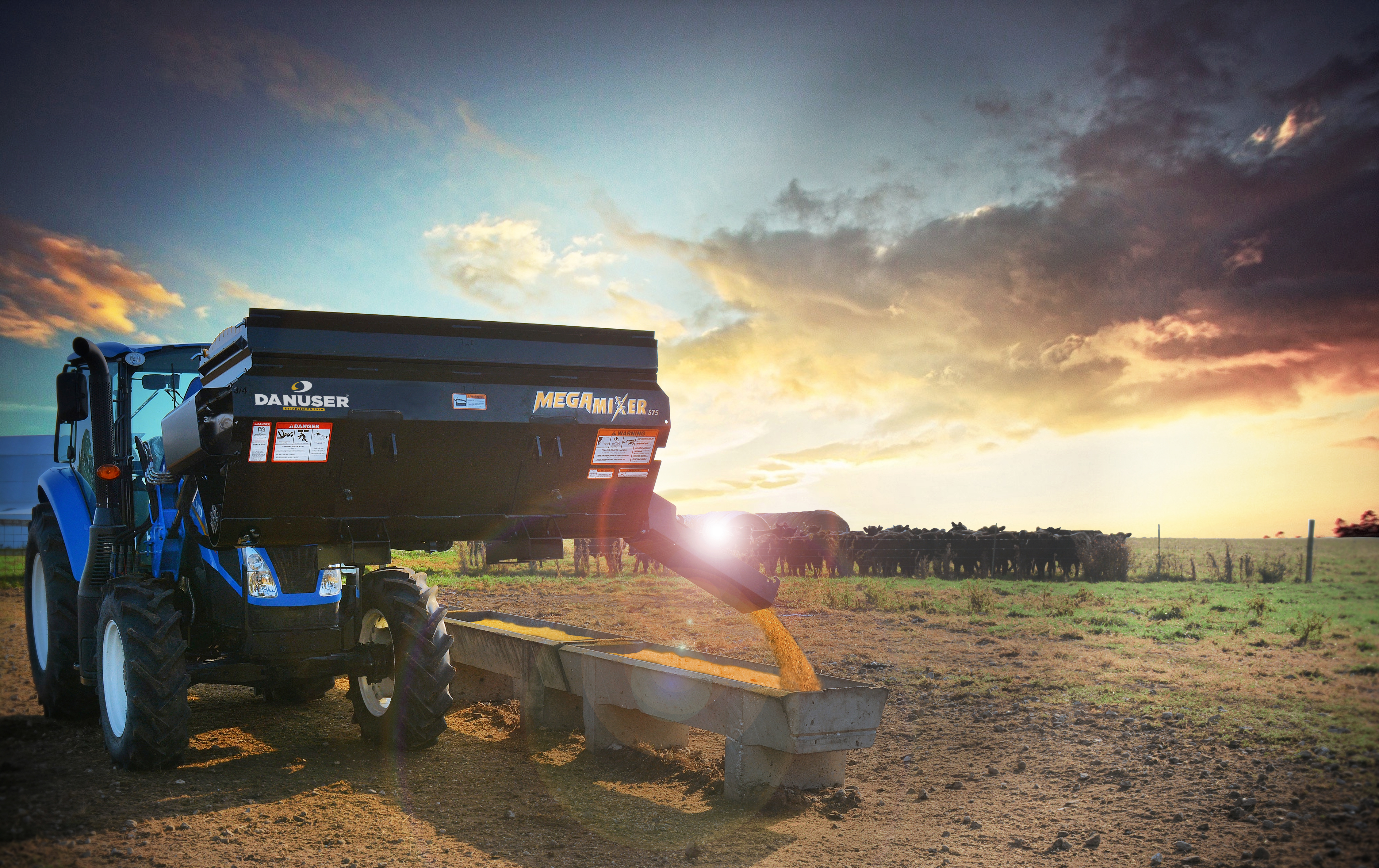 Image showing Danuser Mega Mixer on New Holland tractor pouring feed into trough for cattle under cloudy sky with orange/yellow hue