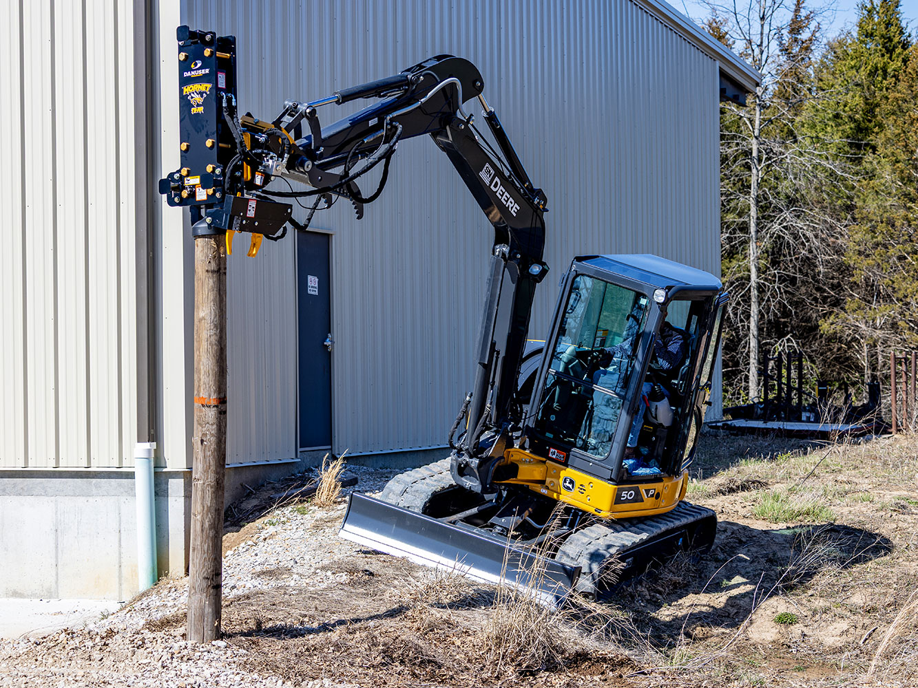 Image showing Danuser Hornet T8-RR post driver on Mini John Deere 50P Excavator holding wood post beside building