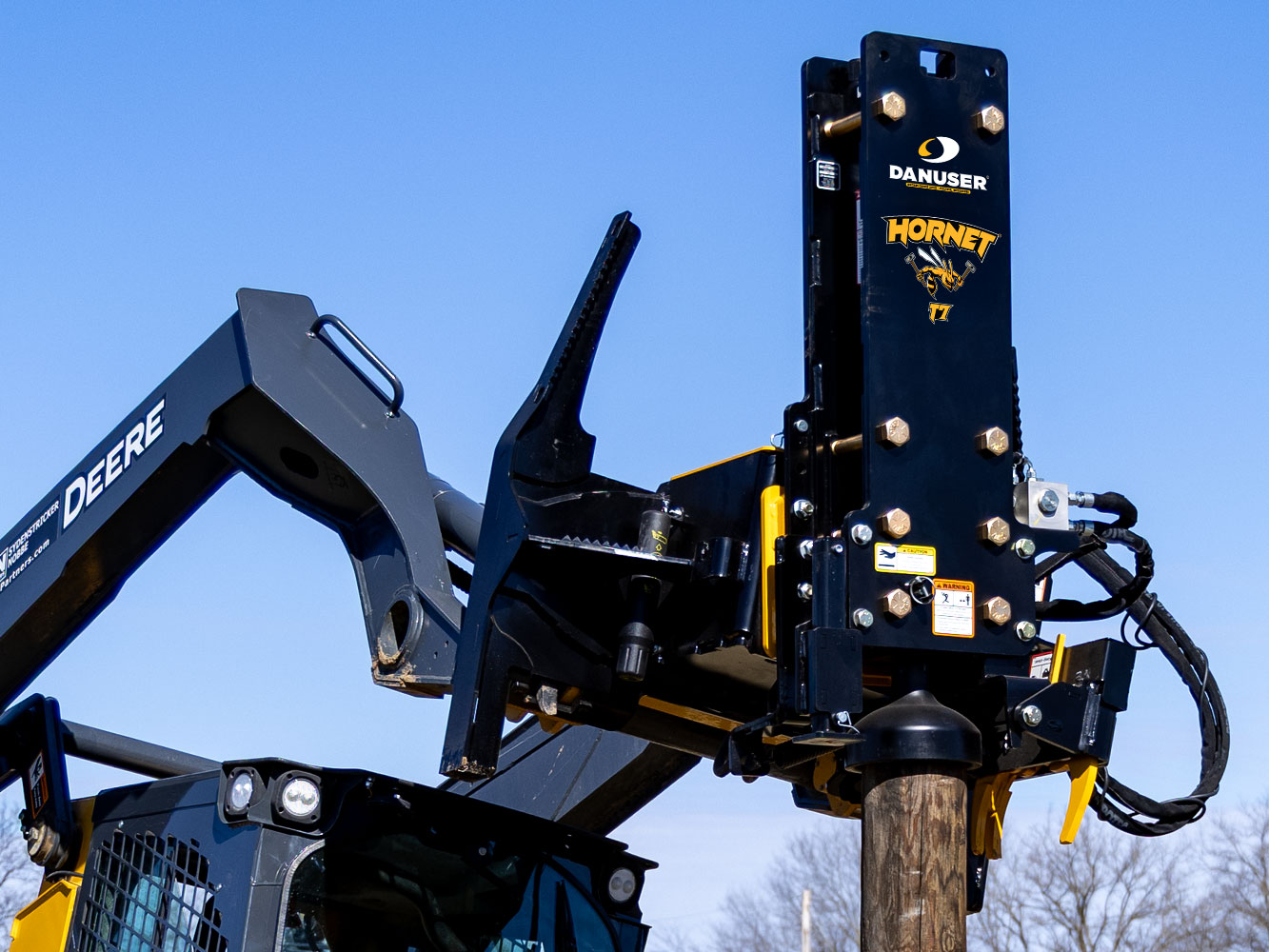 Image showing Danuser Hornet T7 on John Deere 333G Skid Steer shown close-up with sky and leafless trees in the background, holding wood post