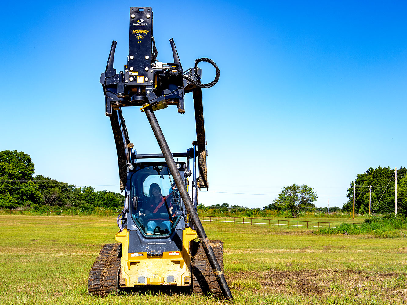 Image shows Danuser Hornet T7 on John Deere 333G Skid Steer using grapple to move post in grassy field for post installation