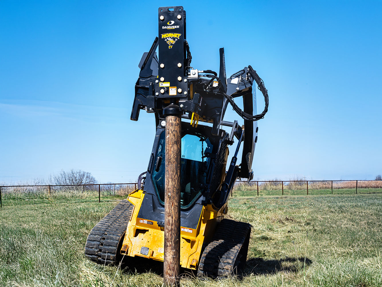 Image showing Danuser Hornet T7 post driver featuring tilt on John Deere 333G Skid Steer driving wooden post into ground in grassy area
