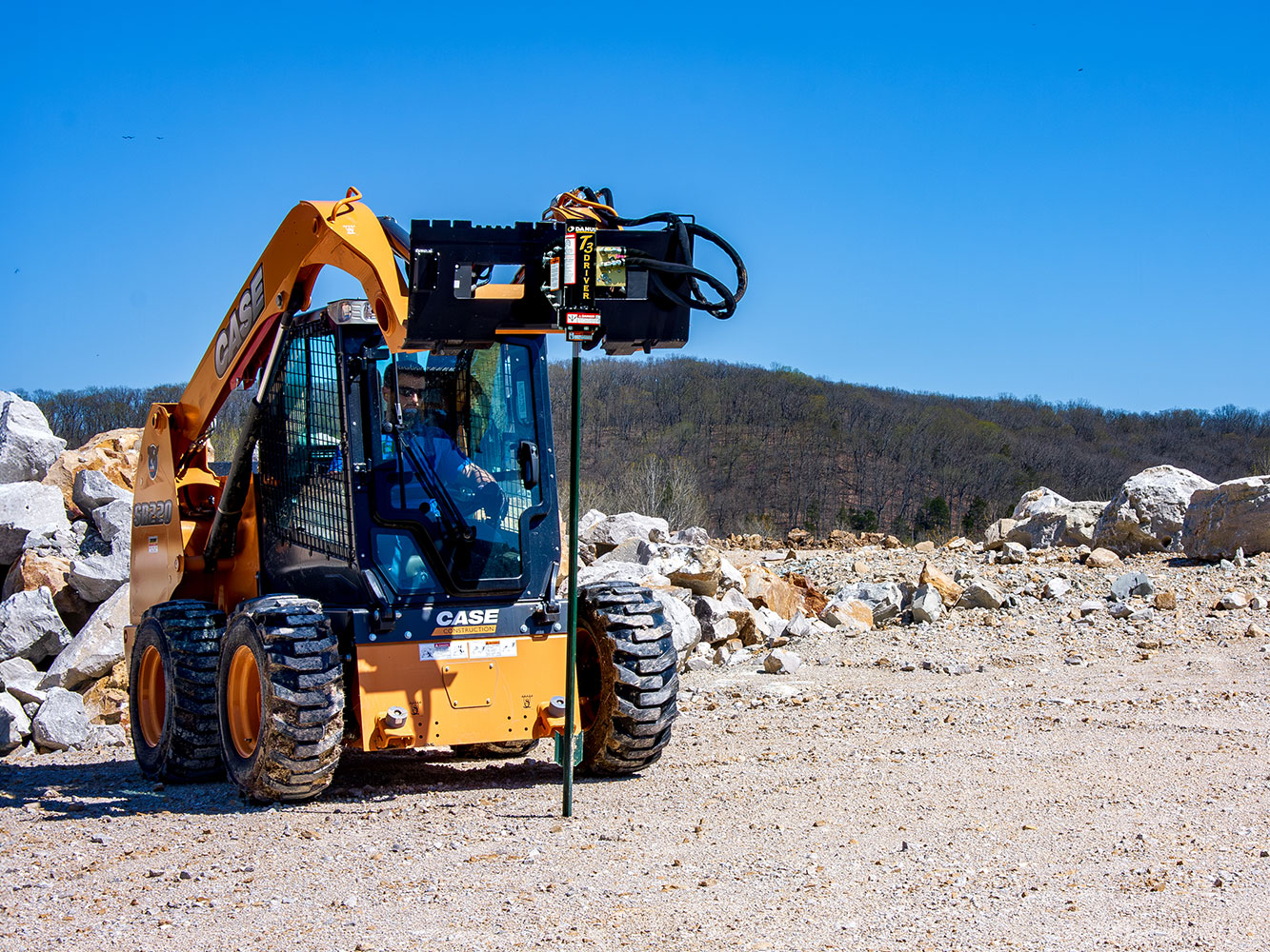 Image shows Danuser T3 post driver on Case SR220 Skid Steer driving T-post into ground in rocky terrain near forested hillside
