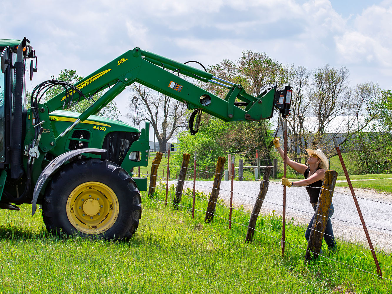 Image shows Danuser T3 post driver on John Deere tractor drives orange T-post as woman holds it steady during fence installation