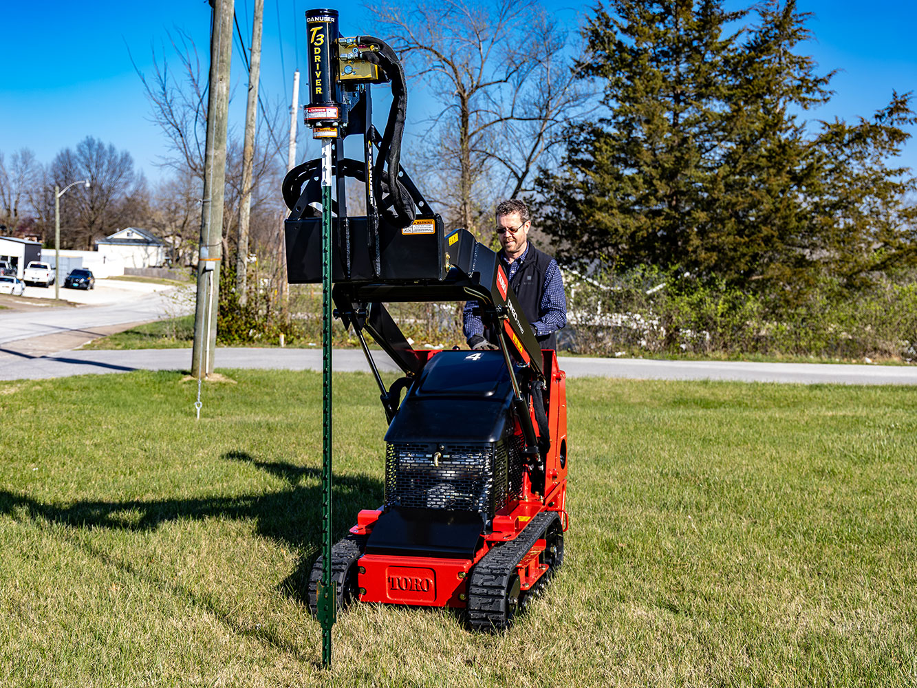 Image of Danuser T3 on red Toro DINGO TX427 Mini Skid Steer driving T-post into ground on grassy area near road