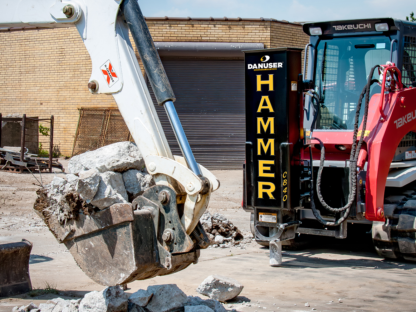 A Takeuchi skid steer with a Danuser Hammer CB40 hydraulic breaker attachment breaks up concrete at a construction site, while an excavator in the foreground lifts debris near a brick building.