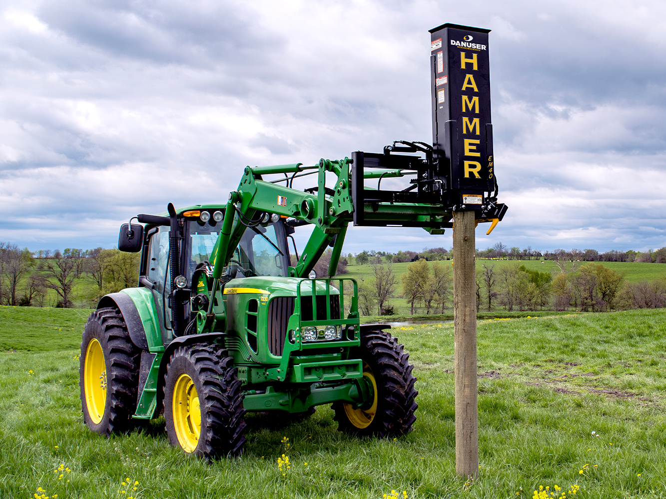 A green tractor with a Danuser Hammer EM40 post driver attachment, driving a wooden post into the ground in an open grassy field with trees and hills in the background under a cloudy sky.