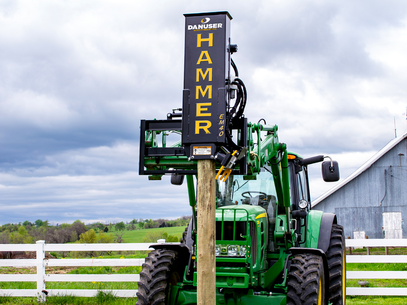 A green tractor with a Danuser Hammer EM40 post driver attachment, driving a wooden post into the ground in a rural setting with a white fence, green fields, and a barn-like structure in the background.