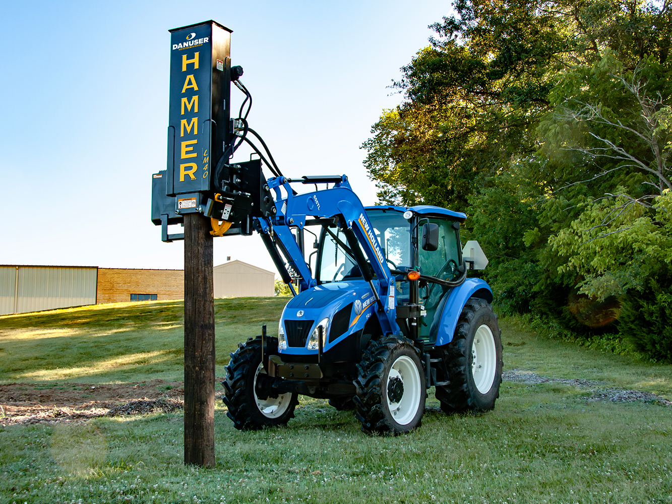 A blue tractor fitted with a Danuser Hammer LM40 post driver attachment, driving a wooden post into the ground in a grassy outdoor area with trees and a building in the background.