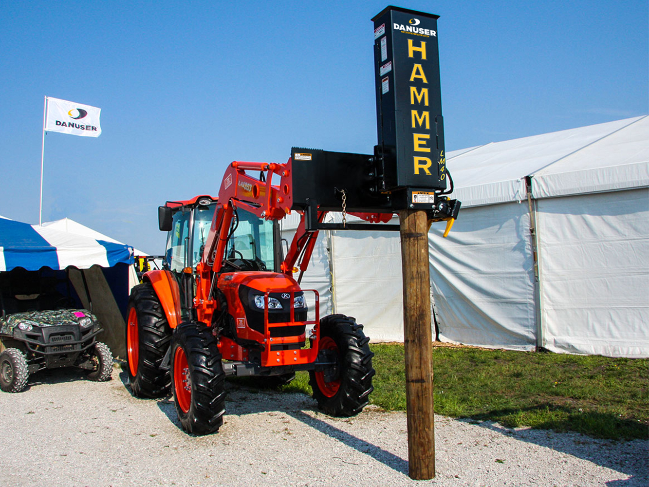A Danuser Hammer LM40 post driver mounted on the front of an orange tractor, driving a wooden post into the ground at an outdoor demonstration area with tents and a Danuser flag in the background.
