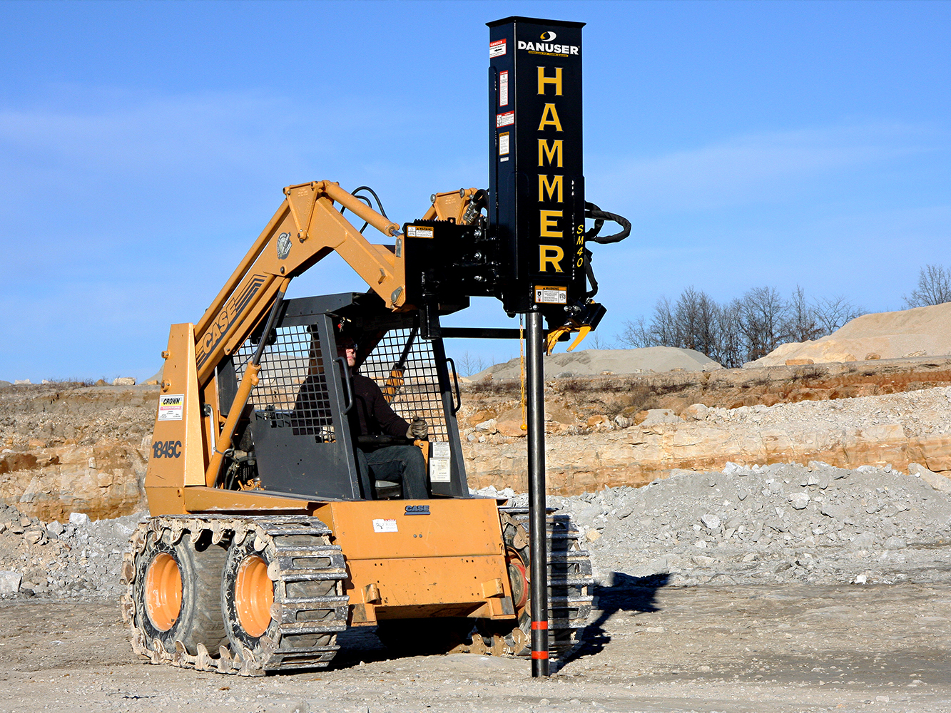 A yellow tracked skid steer loader fitted with a Danuser Hammer SM40 post driver attachment, driving posts into rocky terrain under a clear blue sky, with sparse vegetation in the background.
