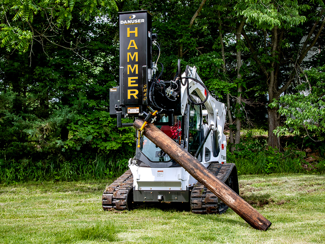 A Danuser Hammer SM40 post driver equipped with a grapple attachment holds a large wooden post, demonstrating its ability to grip and position heavy materials in an outdoor setting.