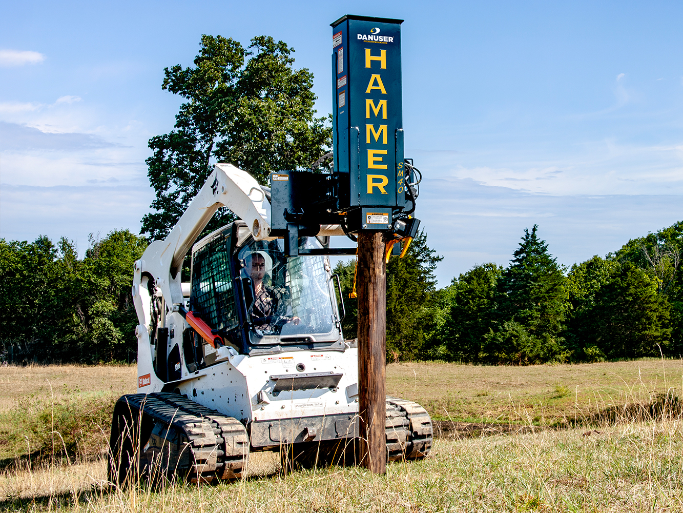 A skid steer loader equipped with a Danuser Hammer SM40 post driver drives a wooden post into the ground in an open field, with trees and a clear sky in the background.