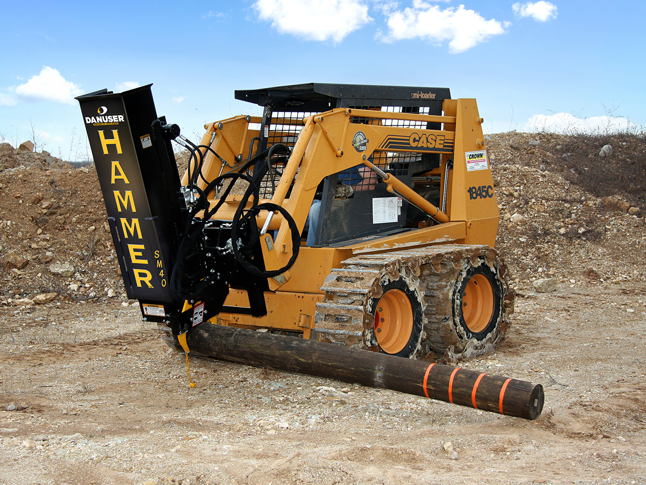 A Hammer SM40 post driver attached to a CASE 1845C skid loader uses its grapple to pick up a large wooden post with orange markings, on a dirt construction site with rocks and a partly cloudy sky.