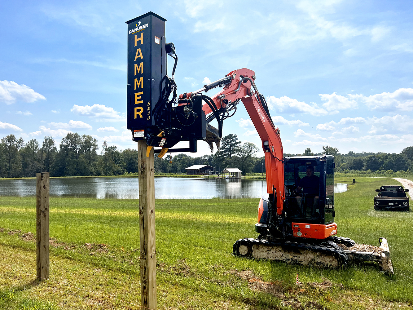 An orange and black mini excavator equipped with a Danuser Hammer SM40 post driver attachment, driving wooden posts into the ground in a grassy area near a pond, with trees and buildings in the background.
