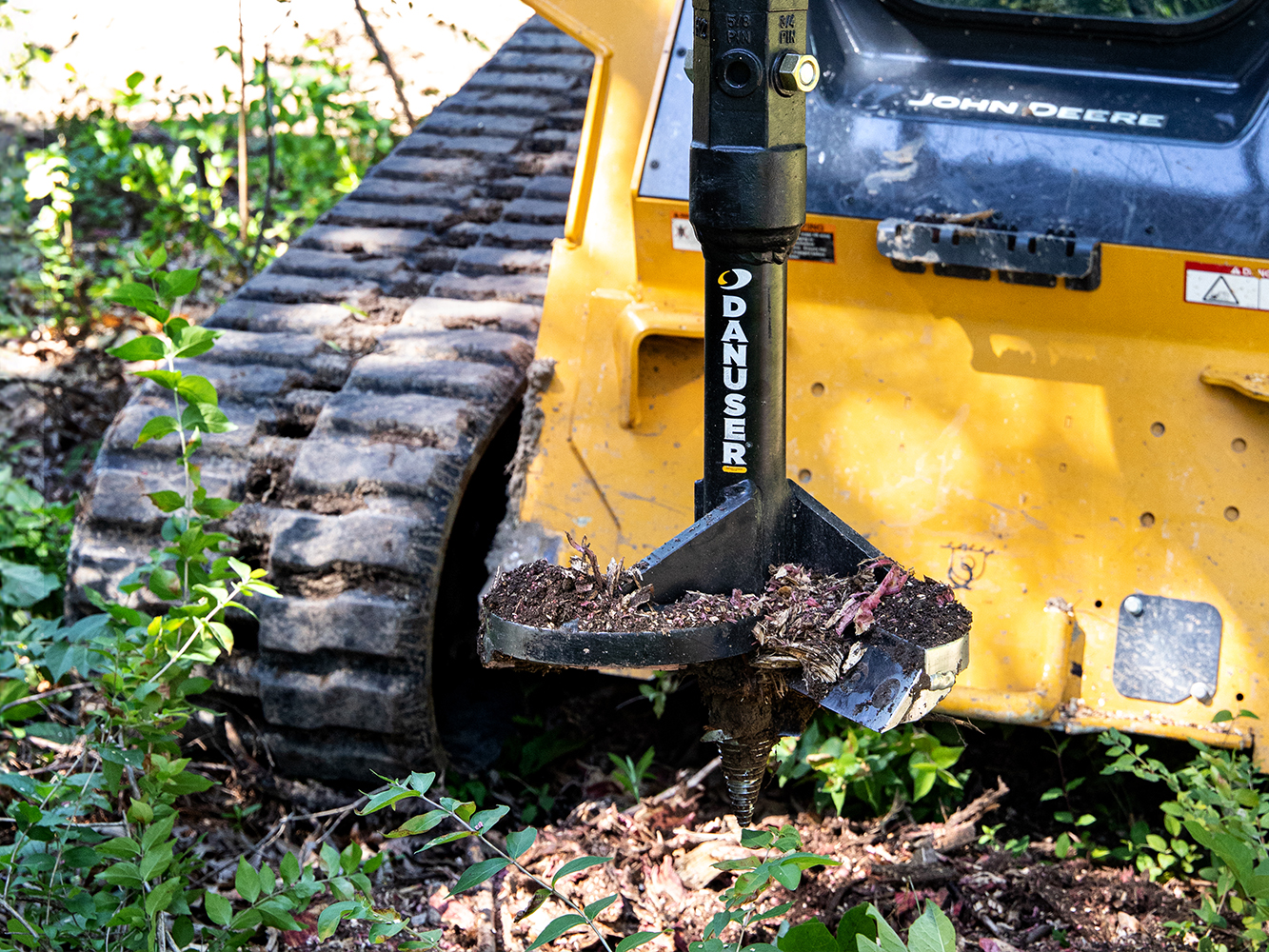 A 16-inch Danuser stump auger attached to a John Deere machine, covered in soil and roots after use, in an outdoor setting with vegetation and dirt, demonstrating its effectiveness in stump removal.