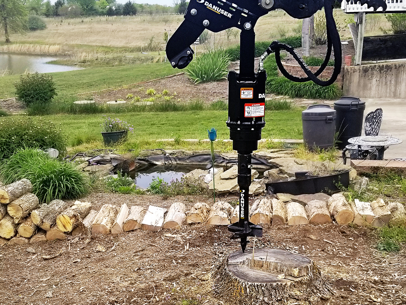 A 10-inch Danuser stump auger attached to heavy machinery, actively drilling into a tree stump in a landscaped yard with cut logs, plants, a pond, and outdoor furniture in the background.