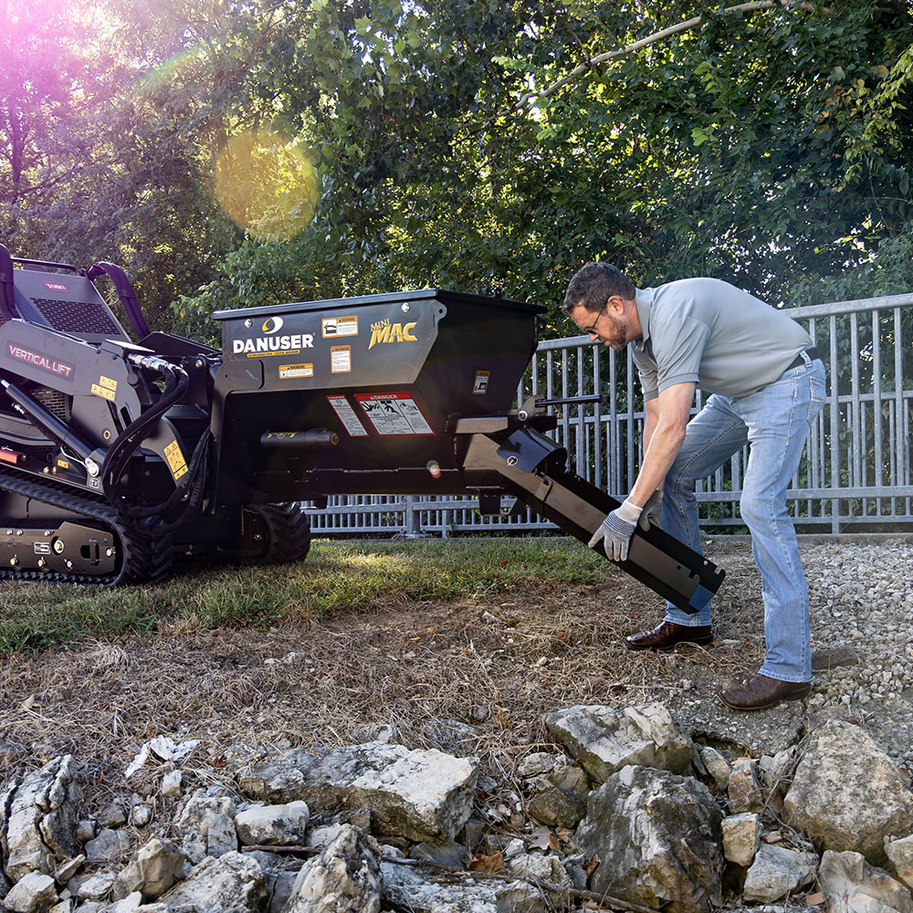 A Danuser Mini Mac material handling bucket mounted on a Toro mini skid steer. An operator, wearing gloves and jeans, is bent down attaching a 24-inch chute to the bucket. The outdoor setting includes trees and a metal fence in the background.