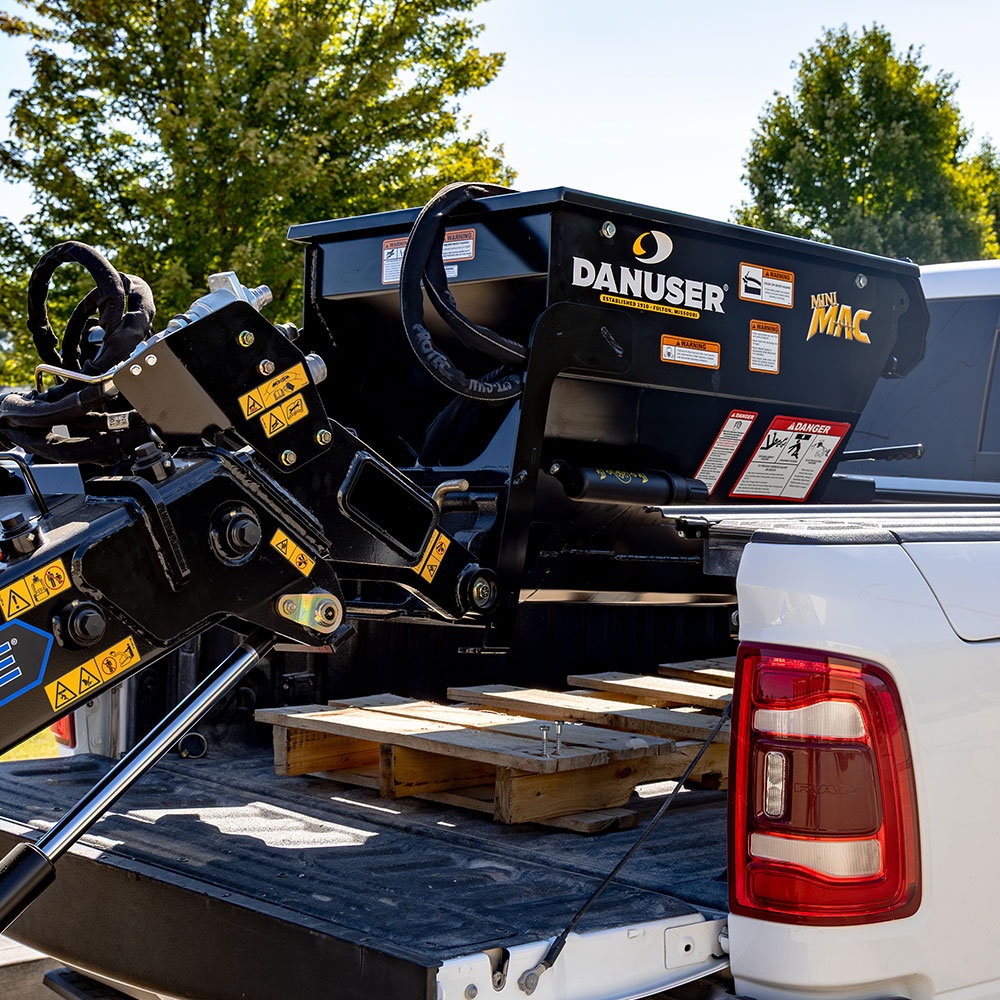 A Danuser Mini Mac material handling bucket being lifted from the bed of a white truck. The black bucket features visible "Mini Mac" and Danuser branding. The truck bed contains wooden pallets, with trees and a clear sky in the background.