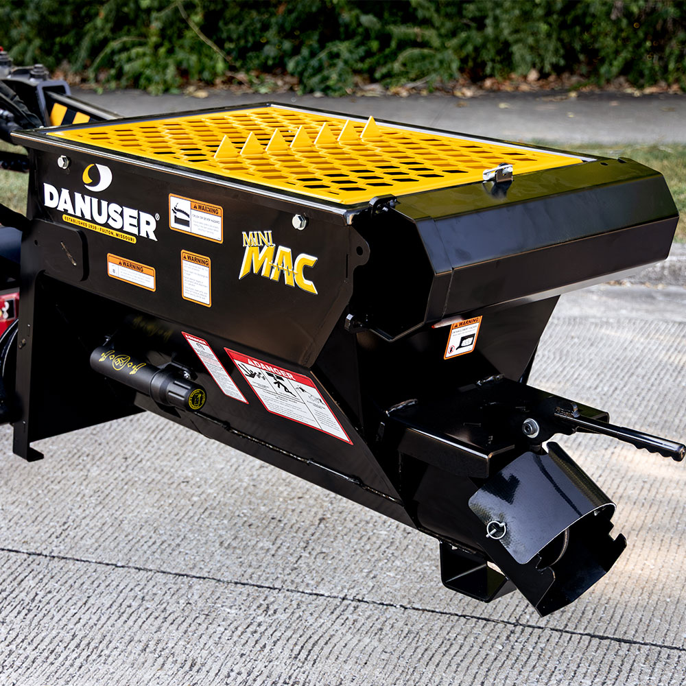 A Danuser Mini Mac material handling bucket featuring a black body with yellow accents, including a yellow guard on top with integrated bag breakers. The side displays the Danuser logo, "Mini Mac" branding, and several warning labels and instructions. The bucket is designed for use with machinery in material handling applications.