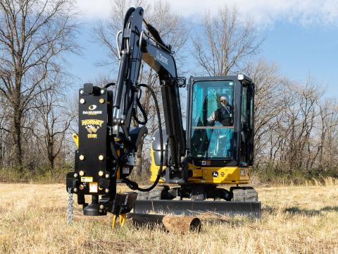 Image showing Danuser Hornet T8-RR post driver on a Mini John Deere 50P Excavator with a wooden post on the ground in a grassy field with trees