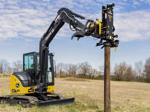 Image showing Danuser Hornet T8-RR post driver on a Mini John Deere 50P Excavator holds a wooden post in a grassy field with trees, fence, and building in the background