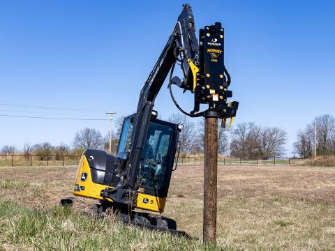 Image shows Danuser Hornet T8-RR post driver with tilt on a Mini John Deere 50P Excavator drives wood post into ground on hillside
