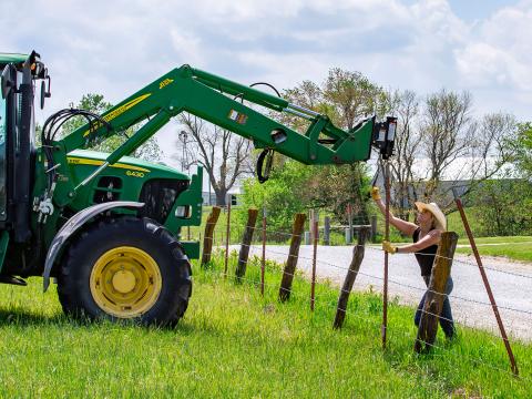 Image shows Danuser T3 on John Deere Tractor driving fence post into ground with woman holding post steady during installation