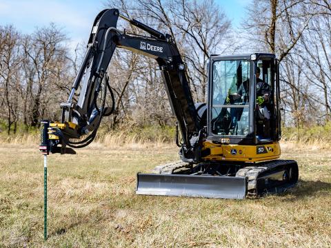 Image shows Danuser T3 post driver on John Deere 50P Mini Excavator driving a post into the ground in a field with trees