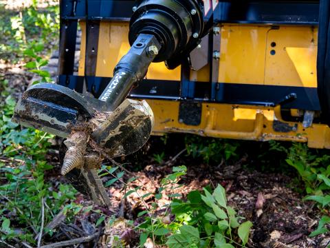 Close-up of a Danuser auger attachment with a large spiral drill bit partially covered in dirt, positioned in an outdoor setting with vegetation and soil, demonstrating its use for ground drilling.