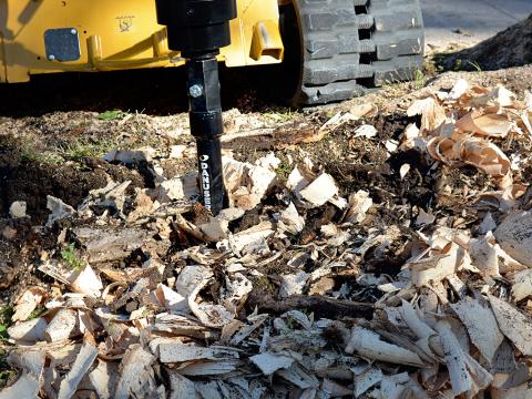 Close-up of a Danuser stump grinder attached to a yellow tracked construction vehicle, actively grinding tree stumps and wood debris, with wood chips and bark scattered across the ground.
