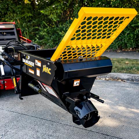 Mini MAC material handling bucket attached to machinery, featuring a black body with yellow accents and a yellow grate on top. Branding includes “Danuser” and “Mini MAC” logos. Positioned on a concrete surface with greenery in the background.