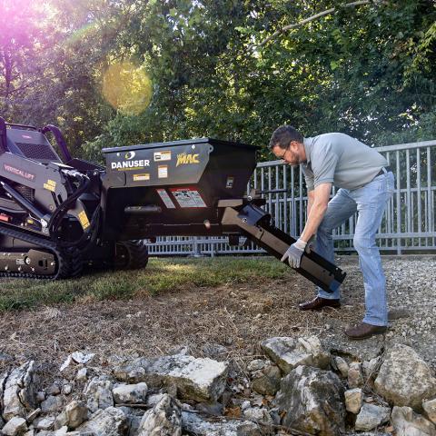 A Danuser Mini Mac material handling bucket mounted on a Toro mini skid steer. An operator, wearing gloves and jeans, is bent down attaching a 24-inch chute to the bucket. The outdoor setting includes trees and a metal fence in the background.