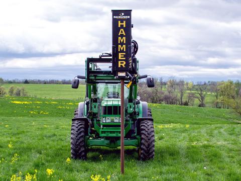 A green tractor in a grassy field with a Danuser EM40 Hammer attachment mounted on the front, used for driving posts into the ground.