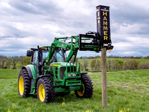 A green tractor using a Danuser EM40 Hammer attachment to drive a wooden post into the ground in a grassy field with rolling hills.