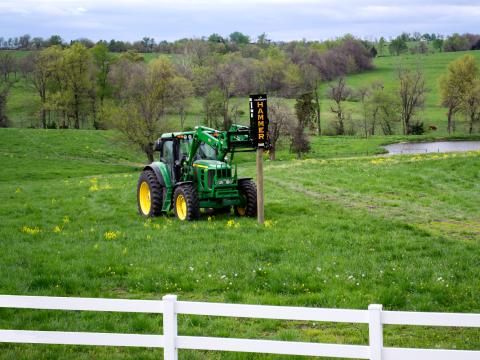 A green tractor with a Danuser EM40 Hammer attachment mounted on the front, parked in a grassy field near a pond and white fence.