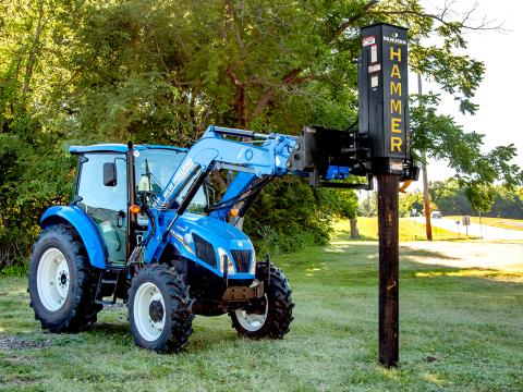 A blue tractor with a front-mounted Danuser LM40 Hammer attachment positioned over a wooden post in a grassy area with trees in the background.