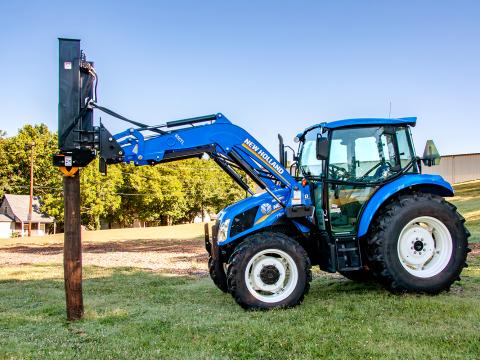 A blue New Holland tractor with a front-mounted Danuser LM40 Hammer attachment lifting a wooden post vertically in a grassy area with trees and buildings in the background.