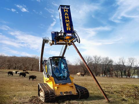 A tracked skid steer loader with a Danuser SM40 Hammer attachment using its grapple feature to load a wooden post into the hammer in a field with grazing cattle.