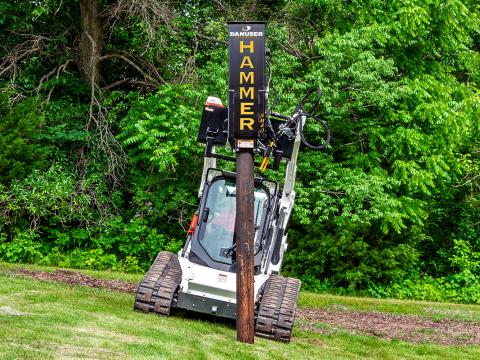 A skid steer loader with a Danuser SM40 Hammer attachment tilted to one side while driving a wooden post into the ground, demonstrating the tilt feature on uneven terrain.