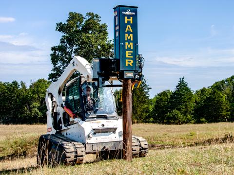 A skid-steer loader with a Danuser SM40 Hammer attachment in an open field, used for driving posts into the ground.