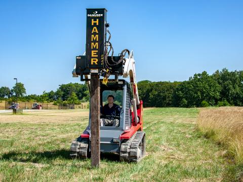 A skid-steer loader with a Danuser SM40 Hammer attachment driving a wooden post into the ground in an open grassy field with trees in the background.