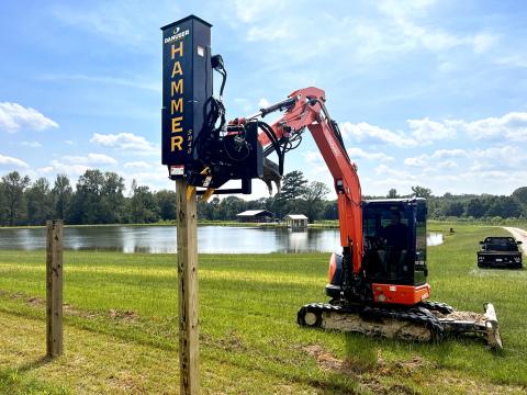 An excavator with a Danuser SM40 Hammer attachment driving a wooden post into the ground near a pond, with trees, buildings, and a vehicle in the background.
