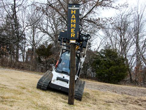 A skid steer loader with a Danuser SM40 Hammer attachment angled to match uneven terrain in a grassy area, using the tilt feature to align a post for driving.