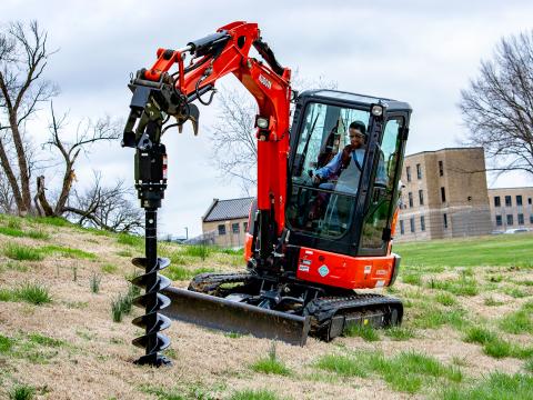 A Kubota KX033‑4 mini excavator equipped with a Danuser EP1020 auger drive, drilling into the ground on a grassy slope with large buildings visible in the background.