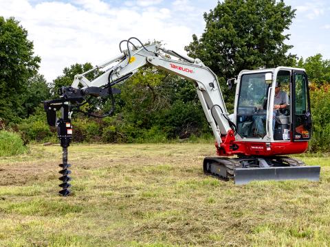 A Takeuchi TB240 mini excavator equipped with a Danuser EP1530 auger drive, drilling into the ground in an open grassy field with trees in the background.