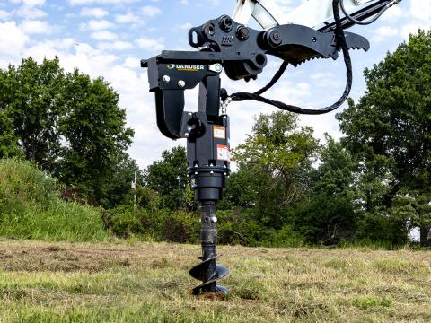 A close‑up view of a Danuser EP1530 auger drive attached to a Takeuchi TB240 mini excavator, with the auger bit mostly drilled into the ground in a grassy field surrounded by trees.