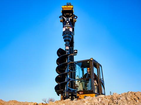 A John Deere mini excavator equipped with a Danuser EP2035 auger drive, drilling into a mound of soil with a large industrial auger under a bright blue sky.